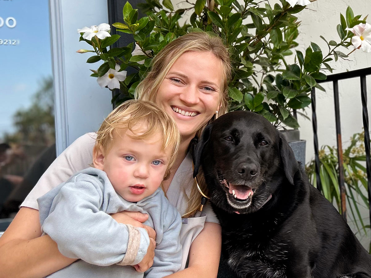 Dr. Zaranek holding a young child and a happy black dog in front of her practice in Pacific Beach